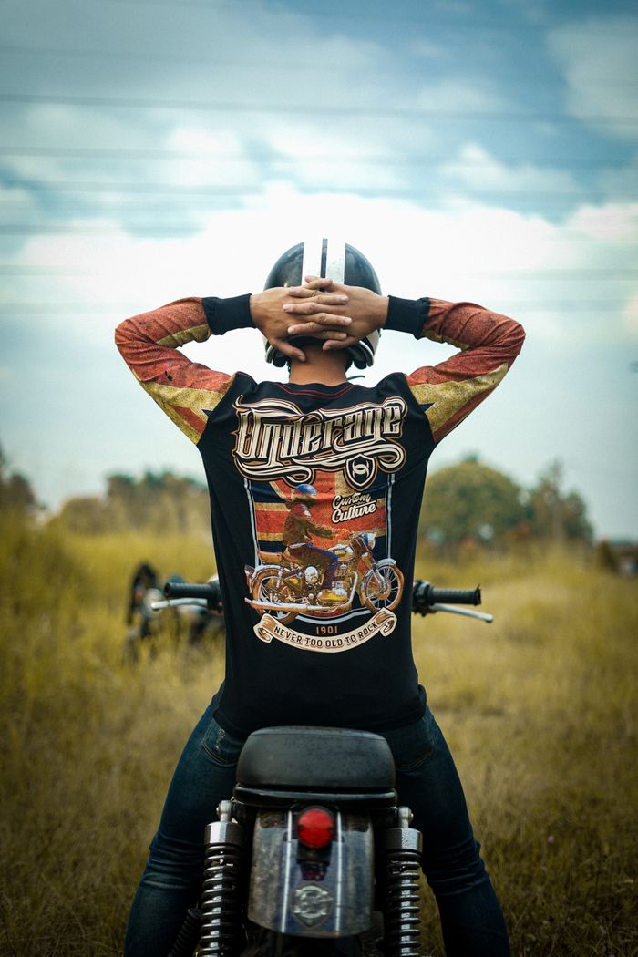 Back view of a biker sitting on a motorcycle in a field, hands on helmet.