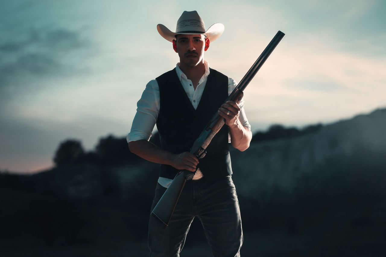 A cowboy in traditional attire holding a shotgun, standing outdoors in San Antonio, Texas.