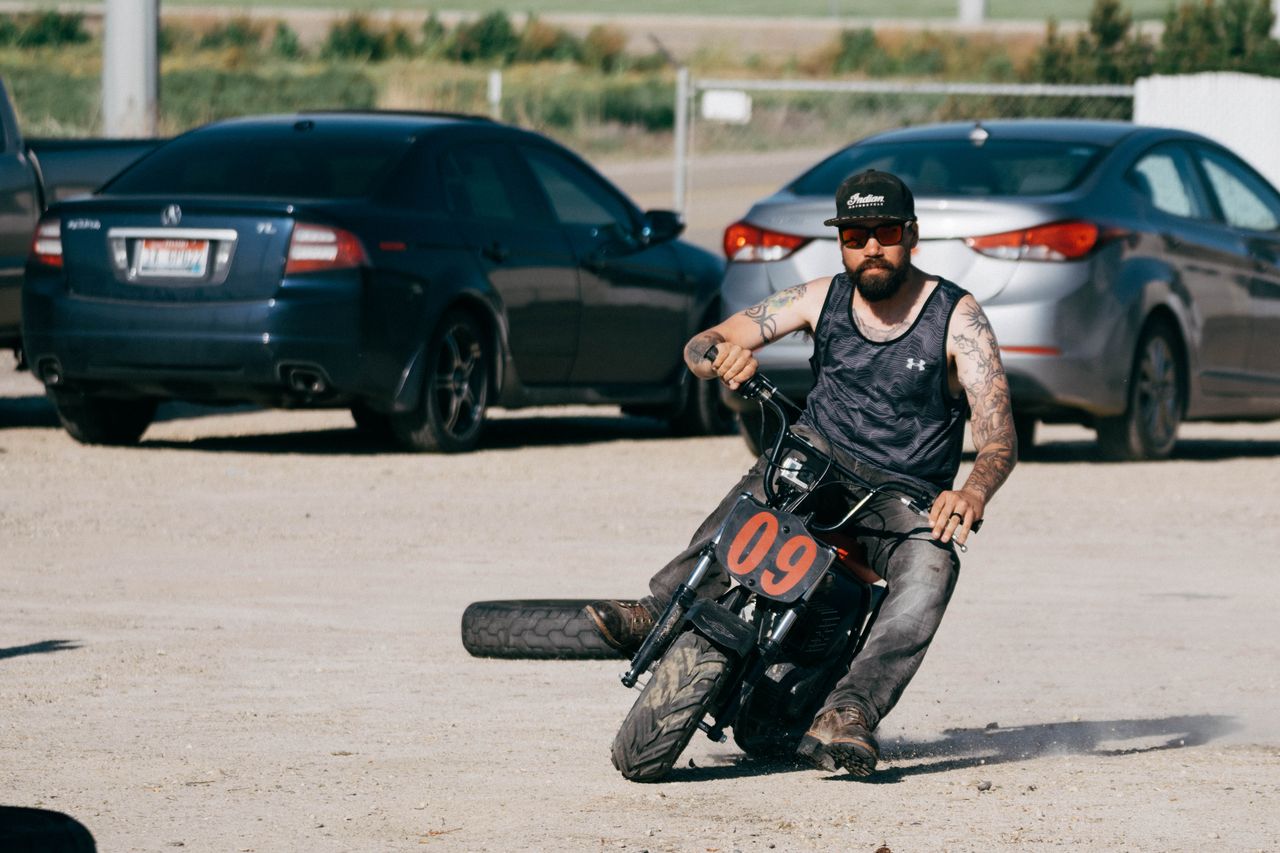 A tattooed man riding a small motorcycle performs a stunt in a sunny parking lot.