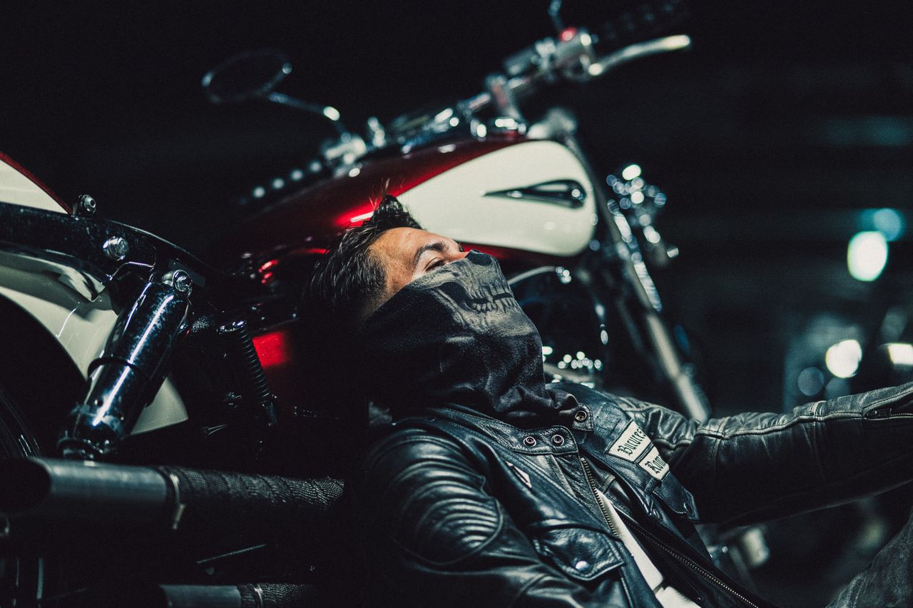 A biker in leather gear rests against a motorcycle under soft lighting with a cool, moody ambiance.